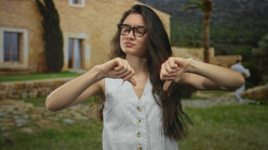 Binanın girişinde duran gözlüklü kadın çıplak kolları ve tarafsız ifadesiyle baş parmak hareketi gösteriyor..