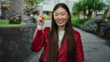 Kırmızı ceketli kadın gündüz vakti şehir sokaklarında gülümsüyor, arkasında ağaçlar ve binalar olan neşeli bir yüz ifadesi sergiliyor..