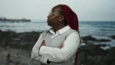 Deniz kenarında kolları çapraz duran bir kadın görünür dalgalarla okyanus ve gökyüzü örgülü saçlarını, kendine güvenen pozunu ve sakin açık hava ortamını vurguluyor..