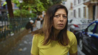 Kadın, şehir caddesinde park halindeki arabalarla ve bulanık binalarla dolu çıplak yüzüyle kameraya bakıyor..