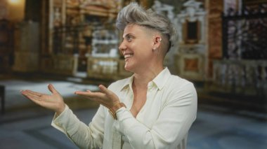 Beyaz tenli, kısa boylu, avuçları açık gülümseyen bir kadın dekoratif bir dini binanın içinde, huzur içinde..