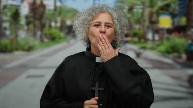 Gri saçlı ve rahip kıyafetli yaşlı bir kadın bir şehir caddesinde duruyor, elinde bir haç, modern bir şehirde maneviyatı ve dini bağlılığı simgeliyor..