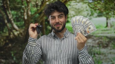 Sakallı genç adam arabanın anahtarlarını tutuyor ve bize dolar veriyor, yemyeşil bir parkta gülümsüyor, açık havada finansal başarı sergiliyor..