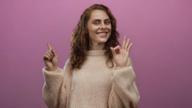 Süveter giyen genç bir kadın pembe bir arka plana işaret ediyor. Yeni stüdyo ortamında pozitiflik ve sadelik taşıyor..