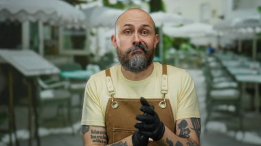 Kahverengi önlüklü kel adam, şehir restoranındaki terasta oturuyor. Açık kafe sahnesinde dövmeli kolları ve değişik yüz ifadelerini gösteriyor.