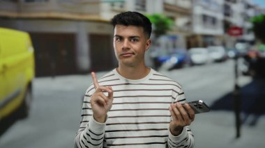 Şehir caddesinde akıllı telefonlu genç bir adam açık havada kargaşa yaratıyor arka planda bulanık araçlar ve binalar olan sıradan çizgili bir gömlek giyiyor..