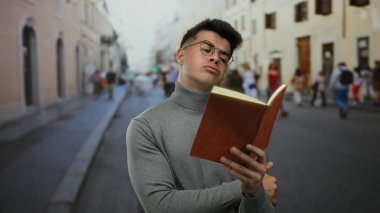 Kalabalık bir şehir caddesinde kitap okuyan genç bir adam arka planda bulanık insanlarla birlikte sayfalara dikkatle odaklanıyor.