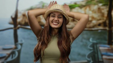 Güneşli bir plajda şapkayla poz veren, deniz kenarında yaz tatili sırasında gençlik neşesi sergileyen bir kadın..