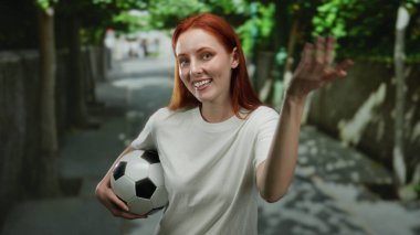 Şehir caddesinde dikilmiş futbol topu tutan bir kadın genç kızıl saçlı güzelliğe odaklanmış canlı şehir manzarasını yakalıyor..
