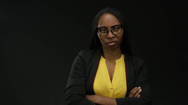 Gözlüklü ve kıvırcık saçlı bir kadın siyah bir duvarın karşısında duruyor. Kollarını çapraz yapıp, sarı bluz ve ceket giyip kararlılığını yansıtıyor..