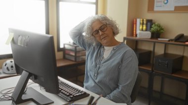 Gri saçlı ve gözlüklü, bir ofis ortamında gerinen bir kadın, bir bilgisayarın önündeki masaya oturmuş, sakin bir iş ortamının örneğini veriyor..