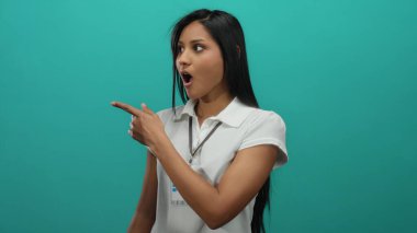 Genç İspanyol kadın, izole edilmiş yeşil bir duvara karşı heyecanla işaret ediyor, beyaz bir gömlek ve kimlik kartı takıyor, çekici bir ifadeyi ve kendinden emin bir hareketi vurguluyor..