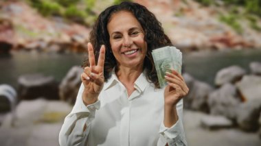 Orta yaşlı bir kadın gülümsüyor, elinde Şili pezosu var ve arka planda kayalar ve su olan deniz kenarında zafer jesti yapıyor..