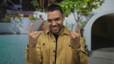 Sarı ceketli adam, güneşli havuz inşaatında iki eliyle kaya boynuzlarını sırıtıyor ve kendinden emin bir şekilde parlıyor..