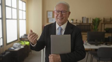 Ofis ortamında dizüstü bilgisayar tutan takım elbiseli kıdemli bir adam kendine güvenen bir ifadeyle profesyonel bir iş ortamı sergiliyor..