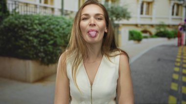 Beyaz elbiseli kadın, güneş ışığıyla aydınlanan binanın önündeki caddede dilini dışarı çıkarıyor; neşeli bir tutum..