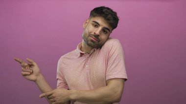 Pembe polo tişört giyen ve pembe bir stüdyoda iki eliyle işaret eden İspanyol genç adam..
