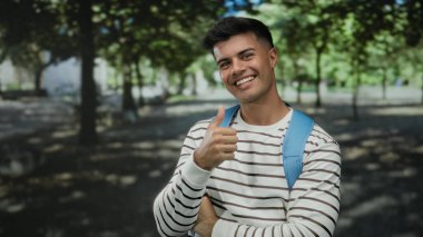 Mavi sırt çantalı genç adam, yemyeşil ağaçlarla çevrili güneşli bir açık hava parkında sıcak bir şekilde gülümserken başparmağını kaldırıyor..