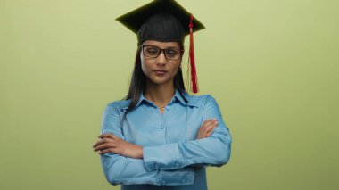 Mavi gömlekli İspanyol bir kadın akademik başarıyı simgeleyen kep ve cübbe giyen parlak sarı bir arka plana karşı ciddi bir ifadeyle poz veriyor..