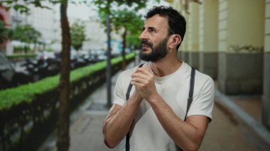 Beyaz tişörtlü İspanyol bir adam şehir caddesinde duruyor. Yeşillik ve park etmiş arabaların yanında düşünceli görünüyor..