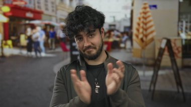 Bulanık insanlar ve arka planda canlı kafeler olan hareketli bir sokakta sakallı genç adam alkışlıyor.