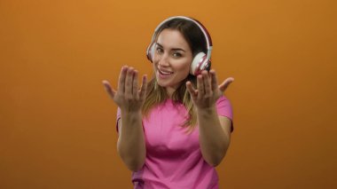 İzole edilmiş turuncu arka planda pembe tişörtlü kulaklık takan bir kadın yüzünde bir gülümsemeyle buraya geliyor..