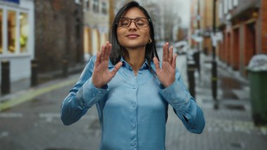 Mavi gözlüklü kadın şehir sokaklarında el kol hareketi yapıyor. Genç ve odaklanmış. Açık şehir ortamında sakin ve anlayışlı..