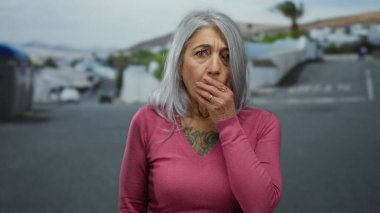 Gri saçlı kadın, arka planda bulanık binalar olan dövmeli göğsünü gösterirken açık bir sokakta pembe bir kazak giyerken ağzını şaşkınlıkla kapatıyor.