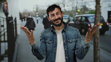 Sakallı İspanyol bir adam kalabalık bir caddede dikiliyor. Kot ceket giyiyor, etrafı trafik ve gezginlerle çevrili, şehir yaşamını somutlaştırıyor ve açık havada günlük zorluklarla boğuşuyor..