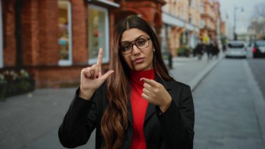 Gözlük takan kadın, şehir sokağında arka planda kırmızı tuğlalı binalarla ezik jestler yapıyor. Dışarıdaki hareketli ortamda kendine güvenen tavırlar sergiliyor..