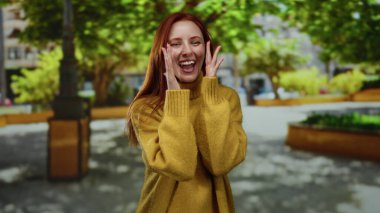 Sıcak bir kazakla gülümseyen bir kadın, canlı bir parkta güneşli bir günün tadını çıkarıyor, yemyeşil ağaçlar ve yeşilliklerle çevrili, bir anlık neşe ve doğal güzelliği yakalıyor..