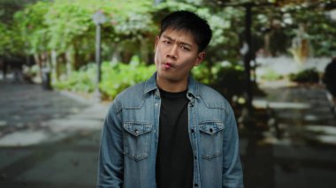 Kot ceket giyen neşeli yüzlü genç adam açık hava parkı ortamında duruyor, arka planda canlı yeşil yapraklarla günlük ve komik bir hava sergiliyor..