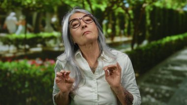 Yeşil bir park ortamında gri saçlı ve gözlüklü yaşlı bir kadın açık havada bulanık yapraklar ve patikalarla çevrili..