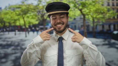 Üniformalı genç İspanyol adam, güneşli bir sokakta yeşil ağaçlar ve bulanık şehir geçmişiyle gülümseyen pozitifliğini ve sıcaklığını iletiyor..