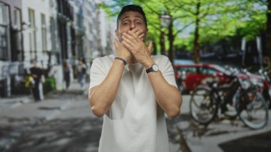 Kalabalık bir caddede park halindeki bisikletler ve kırmızı bir araba, tepesinde ağaçlar ve bulanık yayalar arasında dururken ağzını iki eliyle kapatan bir adam..