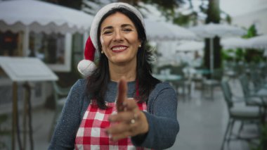 Kırmızı Noel Baba şapkalı ve kareli önlüklü kadın bina cephesinin yanındaki terasta el sıkışmak için elini uzatıyor; misafirperverlik.