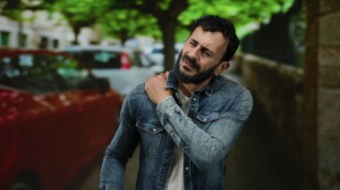 Ağaçları ve arabaları olan, kot ceketli ve sakallı, üzgün bir ifadeyle omzunu tutarak rahatsızlığını gösteren İspanyol bir adam..