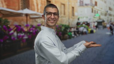 Gözlüklü ve çizgili tişörtlü bir adam canlı bir sokak sahnesinde bulanık kafeler ve renkli çiçeklerle poz veriyor..
