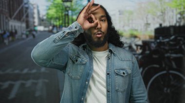 Sakallı bir adam park halindeki bisiklet ve binaların yakınındaki caddede göz göze geliyor. Kot ceket giyiyor. Eğlenceli bir merak..