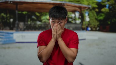 Kırmızı tişörtlü genç adam gün ışığında açık hava parkında şaşırmış bir ifadeyle doğal ortamda çeşitli duygular sergiliyor..