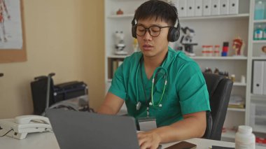 Klinikte ameliyat önlüğü ve kulaklık takan genç bir adam iş ortamında bir dizüstü bilgisayar ve tıbbi ekipman kullanıyor..