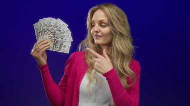 Sarışın genç bir kadın, elinde dolar banknotları tutarken gülümsüyor ve mavi bir arka plana güvenerek finansal başarıyı ve zenginliği sembolize ediyor..
