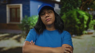 Mavi şapkalı ve gömlekli kadın kurye çapraz kollu ve binanın önünde görünür elleri var; kararlılık.