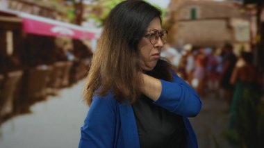 Mavi hırkalı orta yaşlı İspanyol kadın bulanık kalabalığın arasında yoğun şehir caddesinde boynunu tutuyor..