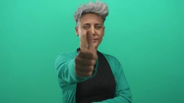 Kısa saçlı bir kadın yeşil stüdyo ortamında yeşil hırka ve siyah bluz giyen bir jest gösteriyor..