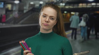 Gözlüklü genç kadın tren istasyonunda renkli kalemler ve çizim defteri tutuyor..