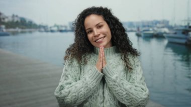 Deniz kenarında sıcacık gülümseyen bir kadın, manzaralı bir limanda rahat bir süveter giyiyor, kayıklar nazikçe yüzüyor, sakin su ve açık hava sükunetiyle çevrili..