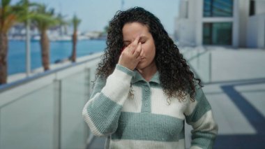 Plaj gezisindeki kadın sıcak süveteri ve okyanus altyapısı ile rahatsızlığını dile getiriyor. Güneşli bir günde palmiyelerin ve modern mimarinin huzurlu manzarasını yakalıyor..