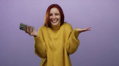 Pürüzsüz mor bir arka planda neşeli bir ifadeyle akıllı telefonu tutan kadın rahat sarı bir kazak giyiyor. Mutluluğu ve modern teknolojiyi temsil ediyor..