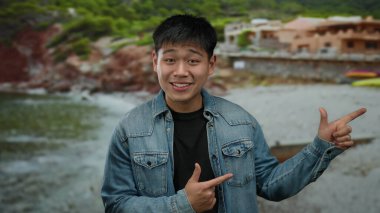 Kot ceketli genç adam arka planda canlı yeşil tepeler ve kırsal evler ile sahil manzarasını coşkuyla işaret ediyor..